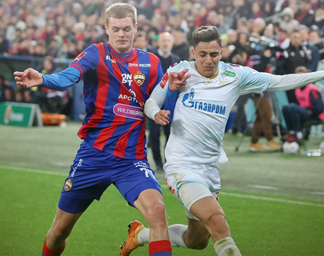 Футболисты в борьбе за мяч на поле. Игрок в красно-синей полосатой форме противостоит сопернику в белой форме. На заднем плане видны зрители и другие игроки