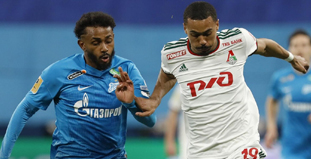 На изображении два футболиста в спортивной борьбе за мяч на поле. Игрок слева в форме синий формы, а игрок справа в белой с зелеными и красными элементами. На заднем плане видны другие игроки и стадион