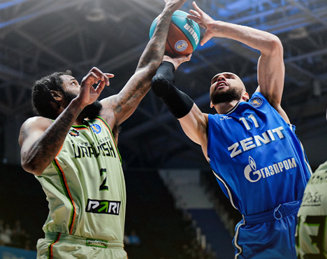 На изображении два баскетболиста в момент борьбы за мяч. Игрок в синей форме с мячом противостоит сопернику в зелёной форме. Действие происходит в спортивном зале